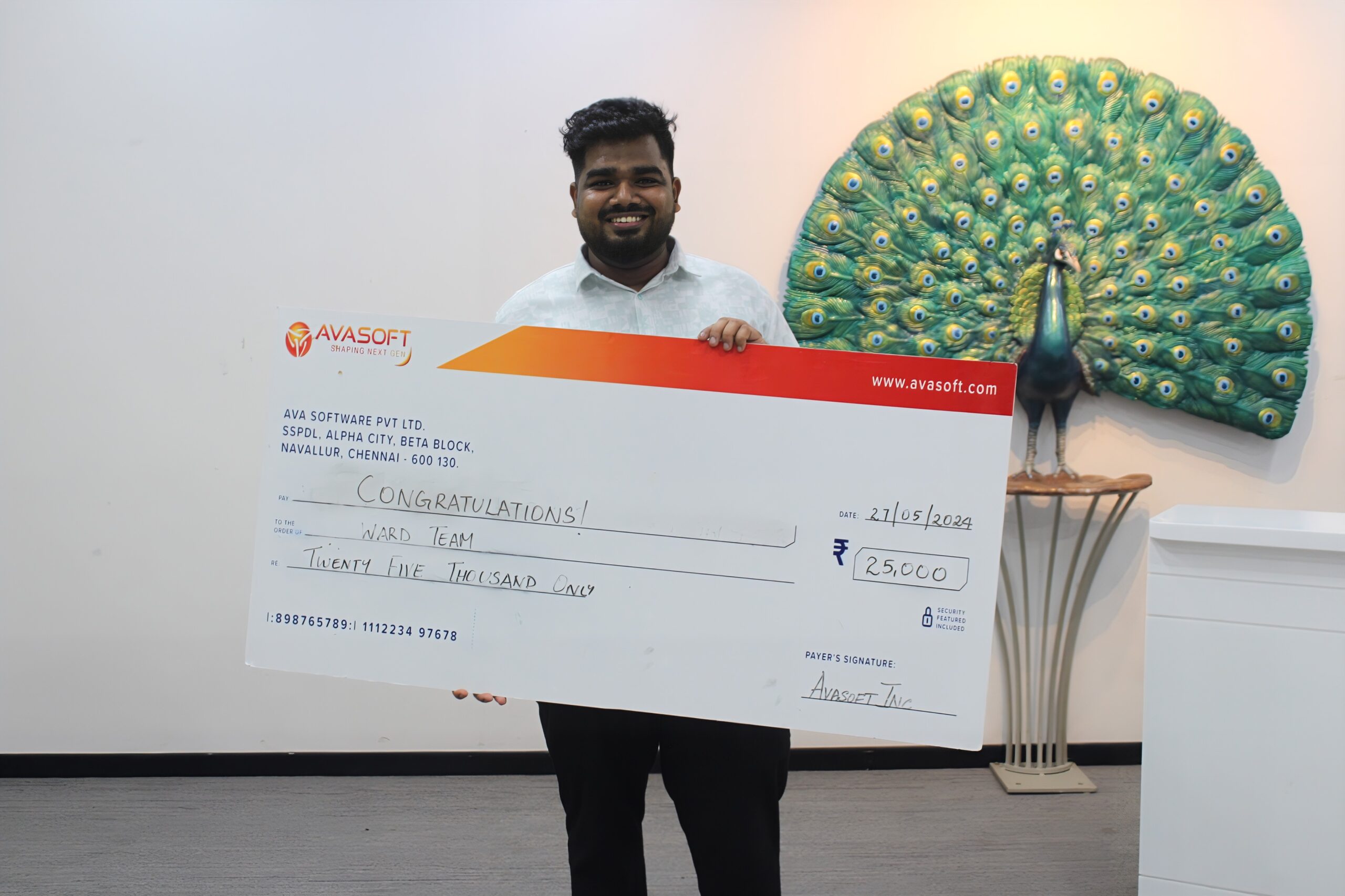 A man is smiling and holding a large check with the amount of ₹250,000, made out to a "Ward Team". The check is from AvaSoft. In the background, there is a decorative peacock artwork displayed on a wall.