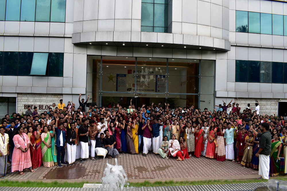 A large group of people, dressed in colorful traditional attire, are gathered in front of a modern office building. They are smiling and waving towards the camera, giving a celebratory feel to the scene. A fountain is visible in the foreground.